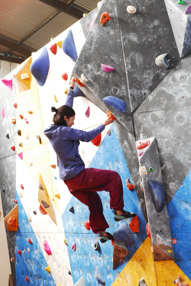 Escaladora con polerón morado y pantalón burdeo en un panel gris y azul, durante una competencia