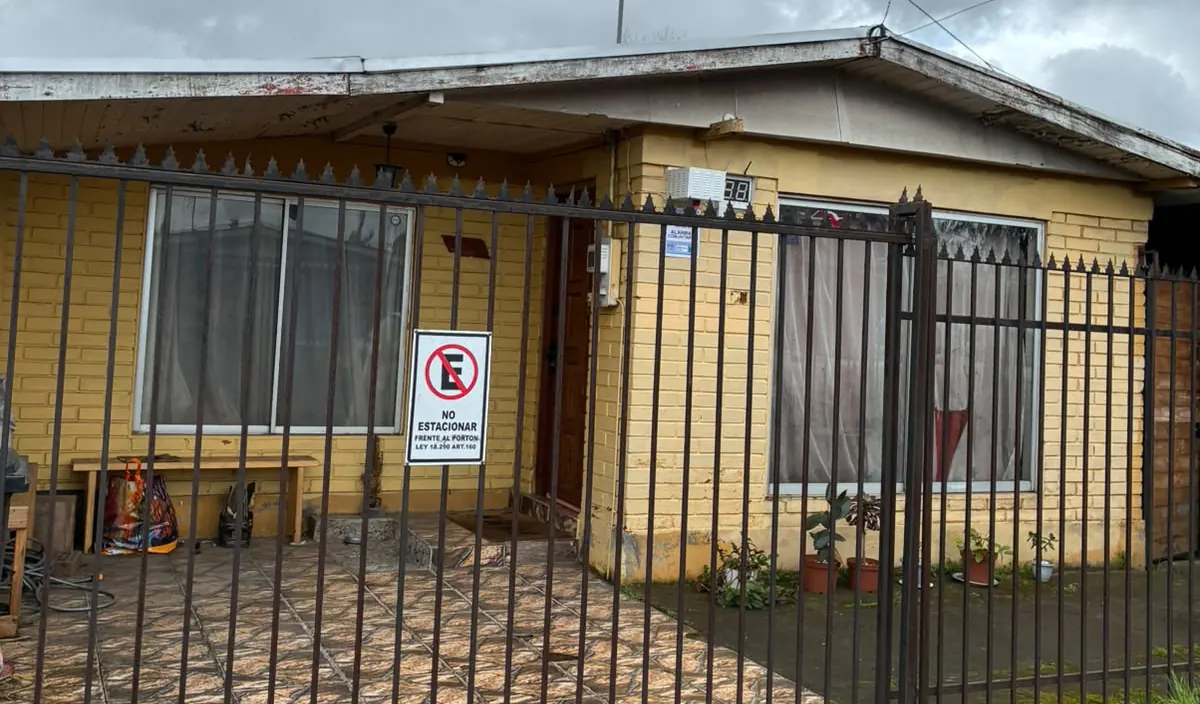 La entrada de Kuyen desde la vereda: la casa de ladrillo amarillo, el portón de reja negra y el pasillo hacia el galpón